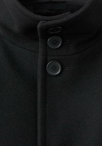 Czarny wełniany płaszcz z wysokim kołnierzem, wyposażony w dwa matowe guziki, prosty fason i gładką teksturę. Zauważalny detal przeszycia.