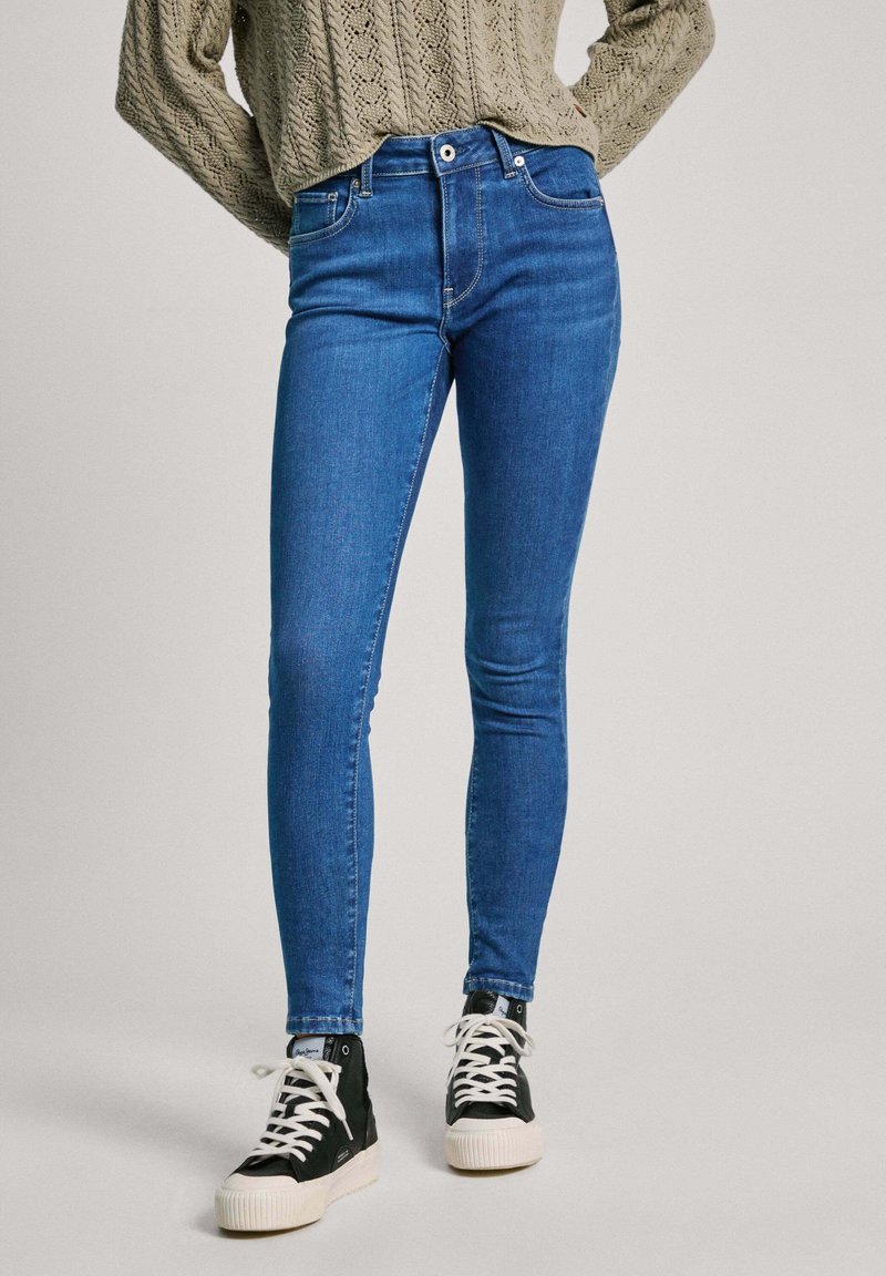 Jeans ajustados de mezclilla azul, con una cintura alta, diseño de cinco bolsillos y un acabado descolorido. Llevados con zapatillas altas negras.