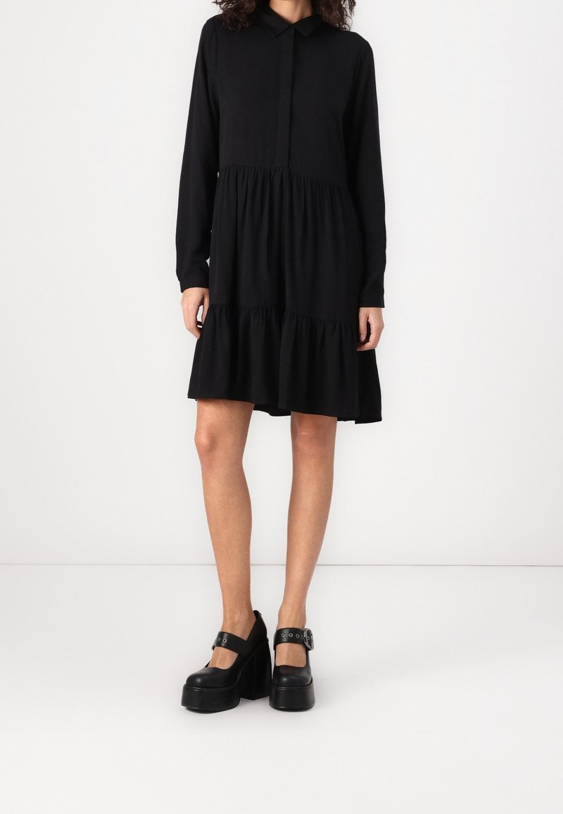 Femme portant une robe noire à manches longues à volants avec un col, et des chaussures Mary Jane noires à plateforme, debout sur fond blanc.