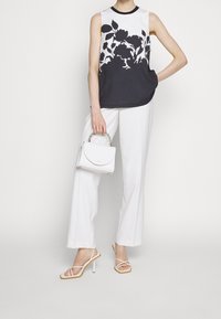 Femme portant un haut sans manches noir et blanc à motifs floraux, un pantalon blanc large, des talons blancs à brides, et tenant un petit sac à main blanc avec une anse en chaîne.