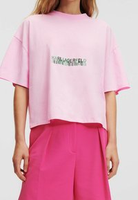 Žena v růžovém zkráceném tričku s textem "Karl Lagerfeld" v zrcadlovém písmu a kontrastními, vysokým pasem a širokými kalhotami v jasně růžové barvě.
