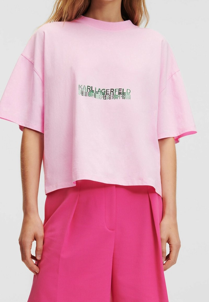 Žena v růžovém zkráceném tričku s textem "Karl Lagerfeld" v zrcadlovém písmu a kontrastními, vysokým pasem a širokými kalhotami v jasně růžové barvě.