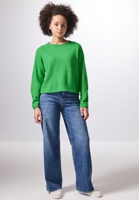 Grön stickad tröja med långa ärmar och rundad halsringning, ihopparad med vida blå denimjeans och vita sneakers. Har en avslappnad passform och textur.