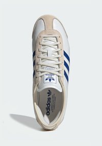 White leather sneaker with beige suede accents, three blue stripes on the side, and a lace-up front. Features a padded tongue and branding.