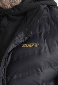 Černá péřová bunda s prošívanou texturou a zipovým uzávěrem. Na prsou je zlaté logo "Siksilk". Mikina pod bundou je černá.