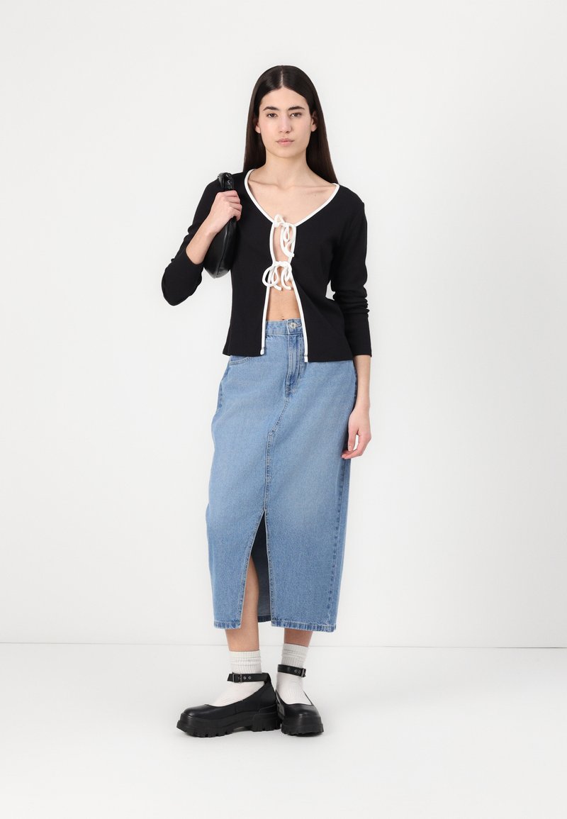 Cardigan nero corto con rifiniture bianche, legato al centro, abbinato a una gonna maxi in denim azzurro chiaro con spacco frontale e scarpe con plateau nere.