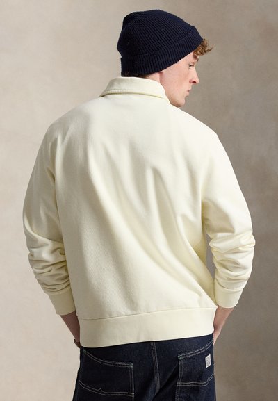 Jeune homme portant un bonnet en maille bleu marine, un pull à manches longues crème et un jean bleu foncé, debout les mains dans les poches arrière, de dos.