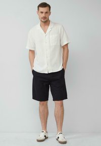 Witte kortgebitte blouse met een zak, gecombineerd met zwarte shorts en witte sneakers met bruine accenten. Glad textuur, casual uitstraling.