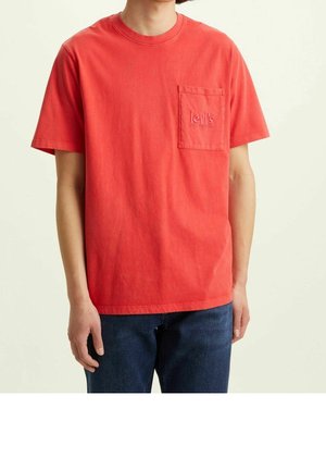 Jeune personne portant un t-shirt rouge à manches courtes avec une poche poitrine et un jean bleu, debout devant un fond clair uni.