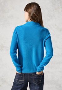 Blauer gerippter Pullover mit hohem Kragen und langen Ärmeln, kombiniert mit dunklen Jeans. Die Textur wirkt weich und gestrickt.