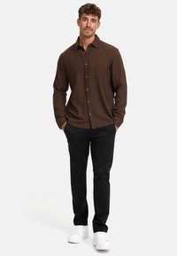 Camisa marrón de manga larga con botones en la parte frontal, combinada con pantalones negros y zapatillas deportivas blancas. Diseño simple, textura suave y ajuste funcional.
