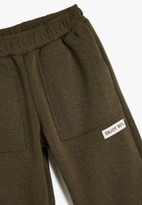 Olivgrüne Jogginghose mit elastischem Bund, zwei seitlichen Taschen und einem strukturierten Stoff. Mit einem weißen Aufnäher mit dem Text "ENJOY 90's".