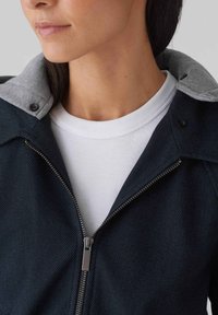 Veste en denim foncé avec un col de hoodie gris clair, détails en fermeture éclair argentée et un t-shirt blanc en dessous. Tissu texturé avec des coutures visibles.