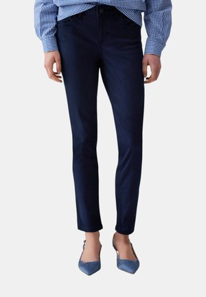 Jeans aderenti blu scuro indossati con una camicia a quadretti blu e bianca e tacchi a punta blu su sfondo chiaro.