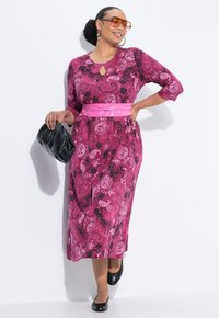Rosa und lila bedrucktes Kleid mit Ausschnitt im Schlüsselaugenstil, Dreiviertelärmeln, schwarzen Akzenten und einem rosa Gürtel. Kombiniert mit schwarzen Accessoires.