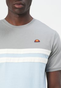 Gray short-sleeve t-shirt with a light blue horizontal stripe and white stripes. Features a small embroidered logo on the chest.
