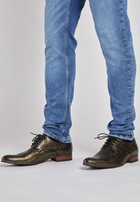 Bronze leather dress shoes with intricate floral embossing, black laces, and a sleek pointed toe, paired with fitted blue jeans.