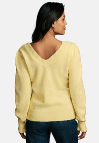 Maglione giallo lavorato a maglia con scollatura a V profonda, maniche lunghe e polsini a coste. Tessuto morbido, vestibilità leggermente ampia, dettagli di cucitura visibili sul collo.