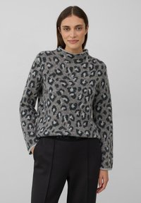 Vrouw in een grijs sweater met luipaardprint en opstaande kraag, met zwarte broek, staande met één hand in de zak tegen een effen achtergrond.