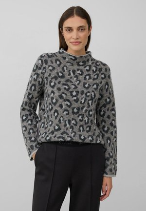 Vrouw in een grijs sweater met luipaardprint en opstaande kraag, met zwarte broek, staande met één hand in de zak tegen een effen achtergrond.