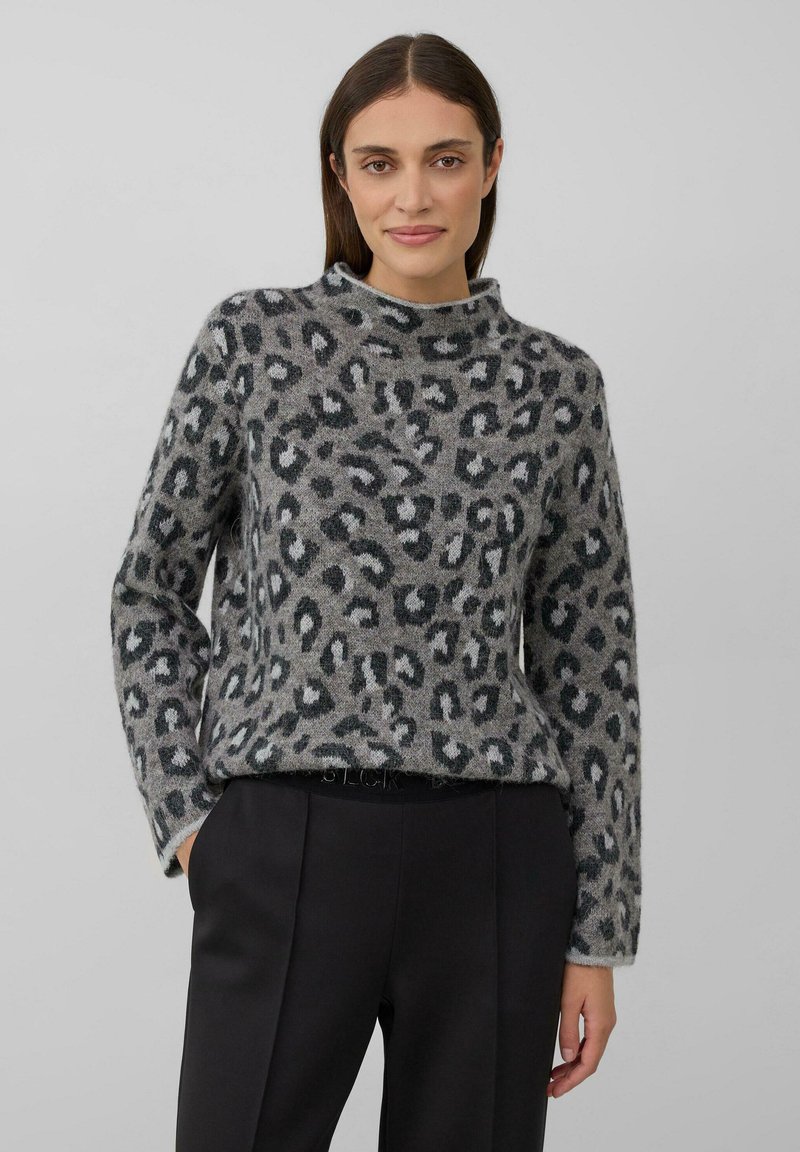 Vrouw in een grijs sweater met luipaardprint en opstaande kraag, met zwarte broek, staande met één hand in de zak tegen een effen achtergrond.