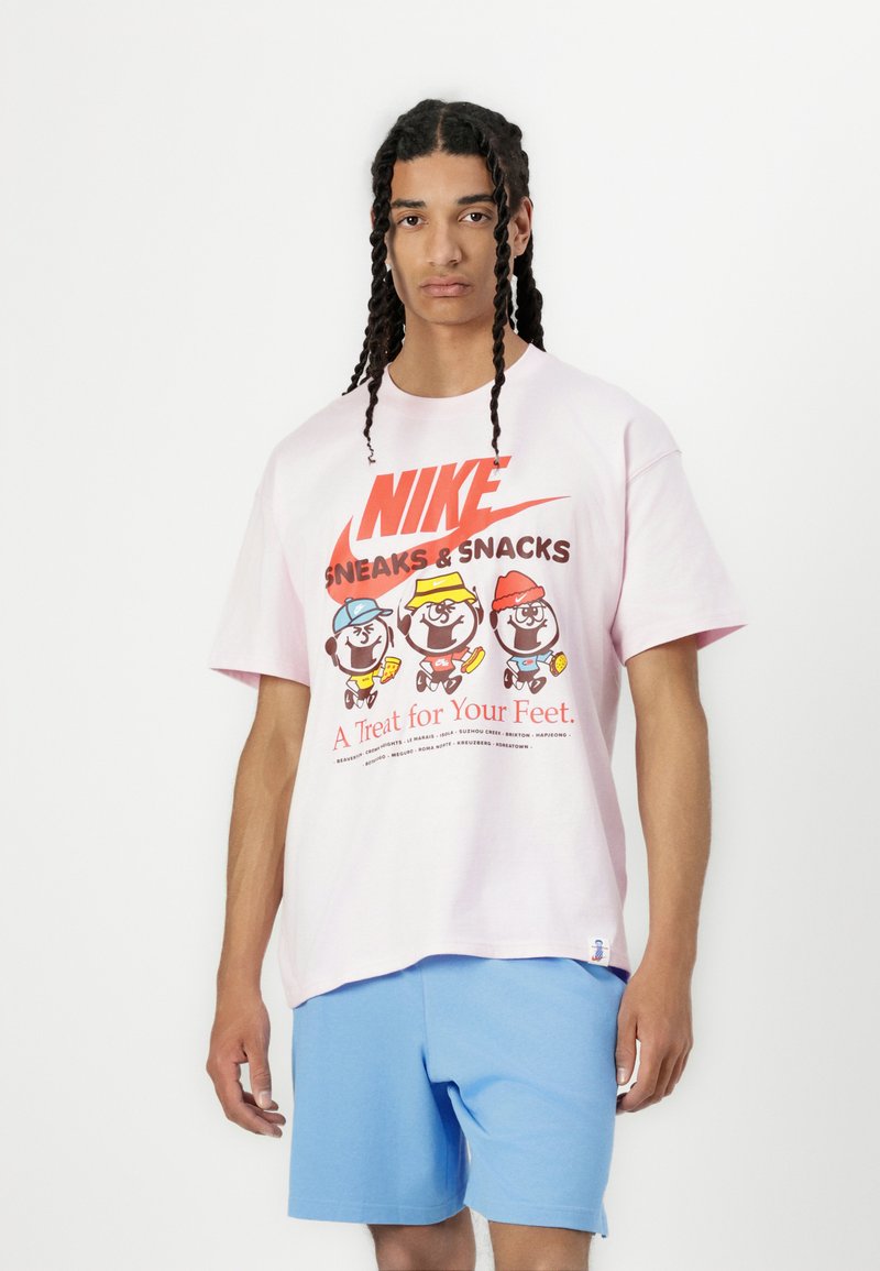 Lyserød bomulds t-shirt med et farverigt grafisk print af tegneseriefigurer med teksten "NIKE Sneaks & Snacks" og blå shorts.
