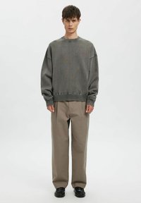 Grauer übergroßer Pullover mit geripptem Kragen und Bündchen, kombiniert mit beigen weit geschnittenen Hosen und schwarzen Schuhen, stehend vor hellem Hintergrund.