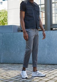 Svart zip-up väst med flera fickor, ihopparad med grå tapered byxor och vita sneakers med svarta och lila detaljer. Utomhusmiljö.