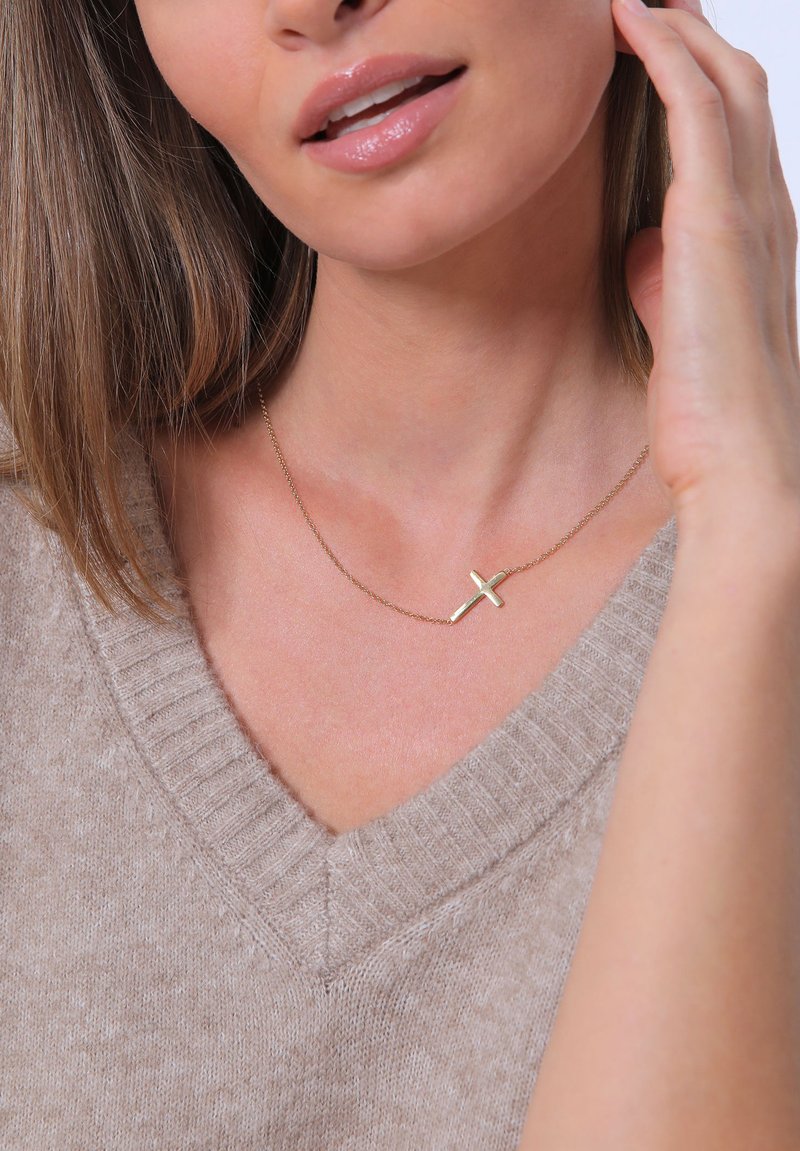 Collana d'oro con pendente a croce minimalista, indossata sopra un maglione beige a scollo a V. Texture liscia, catena delicata e design sobrio.