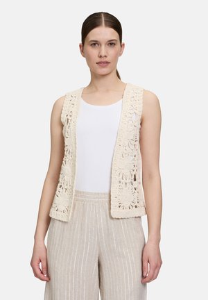 Crochet vest in cream with floral patterns, open front, and no sleeves, paired with light beige striped pants and a white tank top.