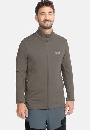 Homme souriant, portant une veste marron zippée Jack Wolfskin et un pantalon bleu-gris avec des renforts noirs, debout devant un fond blanc uni.