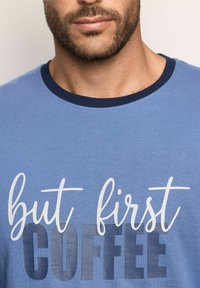 T-shirt in cotone blu con collo rotondo, caratterizzato dalla frase "ma prima COFFEE" in testo bianco e blu scuro. Il testo presenta dimensioni e stili variabili.