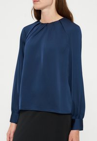 Femme portant un chemisier bleu marine à manches longues avec des plis près du col et une jupe noire sur un fond uni.