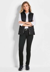 Schwarze gesteppte Weste über einem weißen Hemd mit Kragen, kombiniert mit schmalen schwarzen Jeans und dunklen Sneakers. Einfaches Design mit Frontreißverschluss.