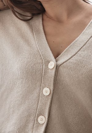 Close-up van een persoon die een beige gebreid vest draagt met drie zichtbare ronde knopen en een V-hals.
