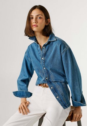 Nuori nainen, joka pukeutunut siniseen farkkupaitaan, jossa on kirjailtuja kuvioita, ja valkoisiin housuihin, istuu puisella jakkaralla yksinkertaisen taustan edessä.