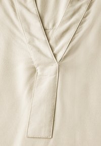 Beige blůza s výstřihem do V, skládacím límcem a jemně texturovanou tkaninou. Čisté švy a hladký povrch.