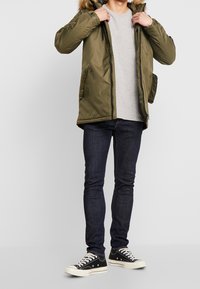 Parka matelassée verte avec capuche bordée de fourrure, deux poches avant, portée sur une chemise gris clair et un jean bleu foncé, associée à des baskets noires.