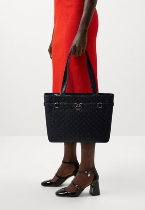Sac tote en cuir verni noir avec motif embossé, deux poignées supérieures et accents en métal doré. Porté avec une robe rouge.