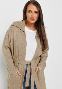 Beige stickad cardigan med sjalkrage, bälte i midjan och ribbad ärm, med två framfickor. Bärs över en vit topp.