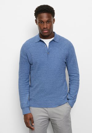 Man draagt een blauw gebreid poloshirt met lange mouwen over een wit ondershirt en lichtgrijze broek, staand met één hand in de zak.