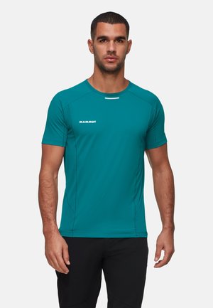 Man in een helder teal sportshirt met korte mouwen en het logo "Mammut" op de borst, gekleed in zwarte broek, staande voor een effen achtergrond.
