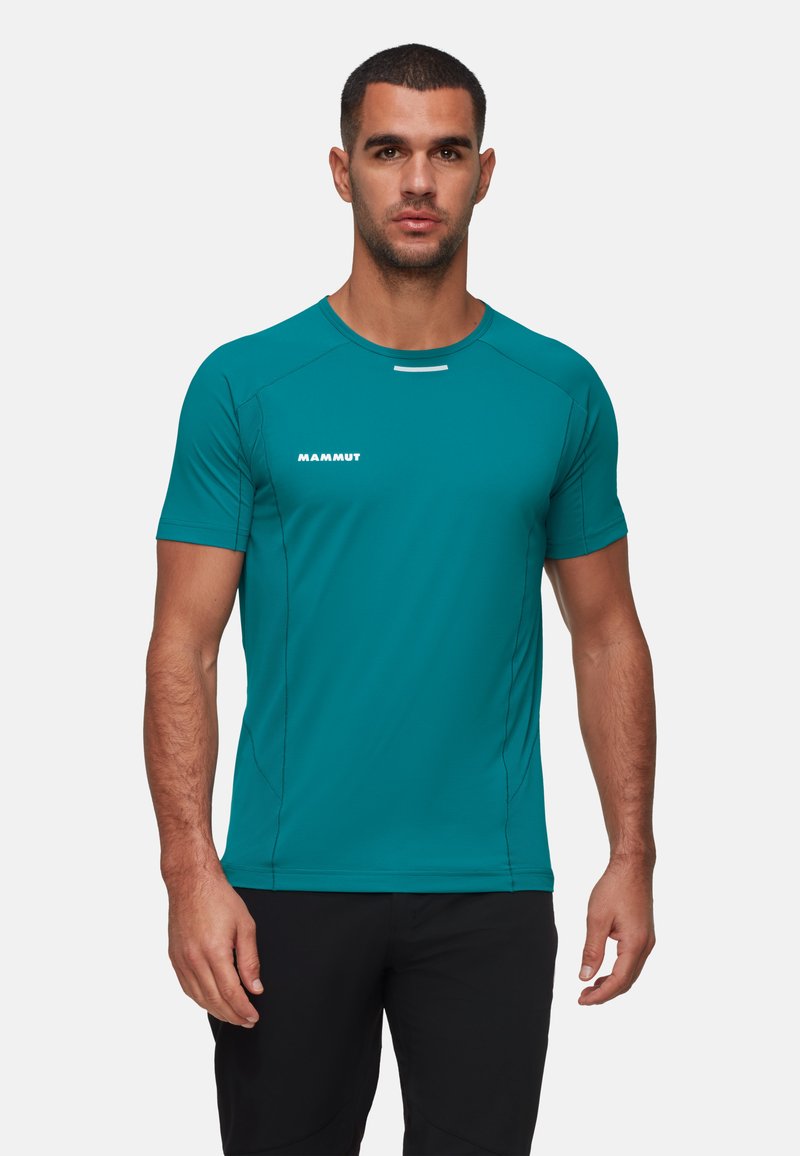 Man som bär en ljus teal kortärmad träningsskjorta med "Mammut"-logotyp på bröstet och svarta byxor, stående mot en enkel bakgrund.