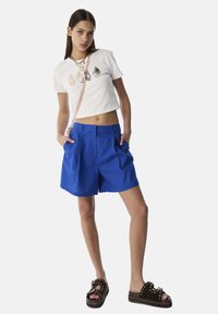 T-shirt blanc court avec des pierres décoratives, associé à un short plissé bleu et des sandales noires avec des accents en forme d'étoile blanche.