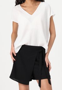 Biały top z dekoltem w serek z lekkiego materiału w połączeniu z czarnymi szortami na zakładkę z wiązanym paskiem w talii; oba elementy mają luźny krój.