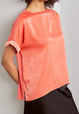 Femme portant un chemisier corail en satin à manches courtes et manches retroussées, associé à une jupe noire, debout contre un mur uni.