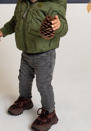 Veste rembourrée verte avec imprimé pingouin, pantalon en denim gris et baskets marron. L'enfant tient un grand cône de pin dans une main.
