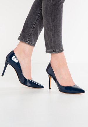 Pieds chaussés de talons aiguilles pointus bleu marine brillants, associés à un jean skinny gris foncé coupé au-dessus de la cheville, debout sur un sol blanc.