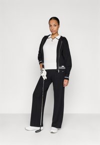 Jachetă neagră din tricot cu motive de cablu, cu accente albe, purtată cu un polo alb și pantaloni negri cu croi larg. Se ține un club de golf în una dintre mâini, în timp ce poartă o mănușă albă.