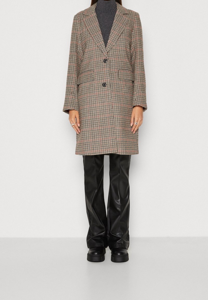 Manteau croisé à carreaux marron et beige, avec des revers crantés et deux poches avant, associé à un pantalon noir en simili cuir.